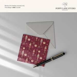May include: A greeting card with a burgundy background featuring a pattern of nutcrackers and gift boxes, with a matching envelope and a black and gold pen. The text "POPPY LANE STUDIO" is visible.