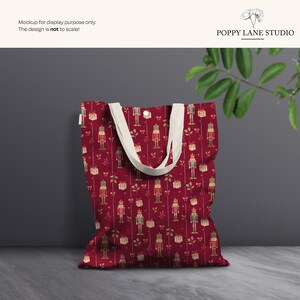 May include: A red tote bag with a repeating pattern of nutcrackers, gifts, and bows in gold. The bag has cream-colored handles and a button closure. The text "POPPY LANE STUDIO" is at the top.