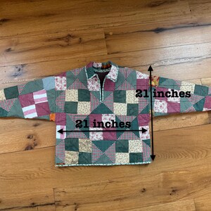 May include: A long-sleeved pullover shirt made of quilted fabric with a patchwork pattern in shades of green, red, and cream. The shirt features a collar and a partial front zipper. Measurements indicate a width and length of 21 inches.