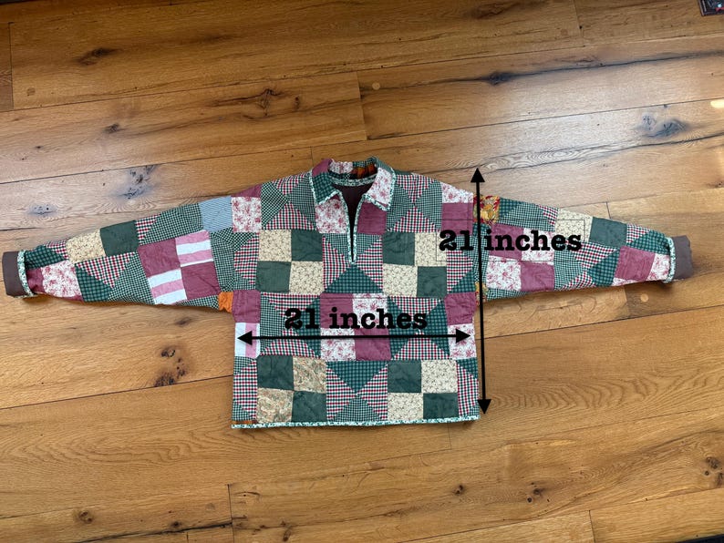 May include: A long-sleeved pullover shirt made of quilted fabric with a patchwork pattern in shades of green, red, and cream. The shirt features a collar and a partial front zipper. Measurements indicate a width and length of 21 inches.