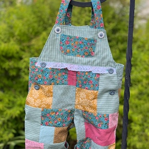 May include: A patchwork baby overall with floral and striped patterns in shades of green, pink, and yellow. The bib overall has a pocket, daisy-shaped buttons, and white lace trim. The garment is hanging from a black metal bar.