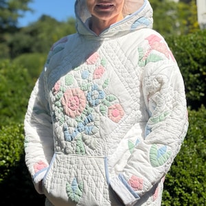 May include: A white quilted hooded sweatshirt featuring pastel pink, blue, and green floral appliqué patterns. The garment includes a front kangaroo pocket, long sleeves, and light blue trim along the hood, cuffs, and pocket edges with diamond stitching.