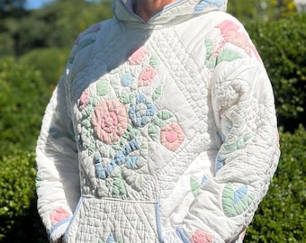 May include: A white quilted hooded sweatshirt featuring pastel pink, blue, and green floral appliqué patterns. The garment includes a front kangaroo pocket, long sleeves, and light blue trim along the hood, cuffs, and pocket edges with diamond stitching.