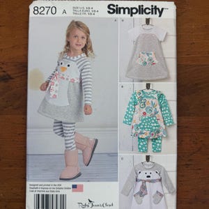 May include: Simplicity sewing pattern 8270 for sizes 1/2-4. The envelope features illustrations of dresses and leggings, including a penguin-themed dress, a floral print dress with matching leggings, and a polar bear-themed dress with a scarf.