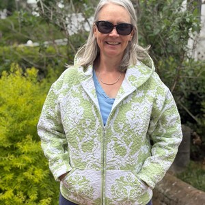 May include: A quilted zip-up hooded jacket featuring a light green and white damask floral pattern. The garment includes a full-length front zipper, long sleeves, and two front patch pockets. The hood is attached to the collar area.