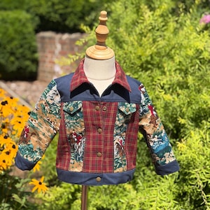 May include: A long-sleeve jacket featuring a mix of navy blue solid fabric, red and green plaid panels, and a printed fabric depicting equestrian scenes with riders on horses. The jacket has a pointed collar, button-front closure, and two chest pockets.
