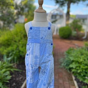 May include: Light blue patchwork overalls with floral and plaid patterns. The overalls feature blue buttons on the straps and sides. The garment is displayed on a mannequin in an outdoor setting, showcasing a vintage style.