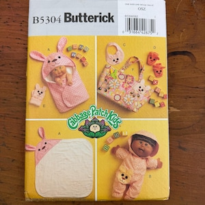 May include: A Butterick sewing pattern for Cabbage Patch Kids doll clothes and accessories. The pattern features a bunny-themed hooded blanket, bib, tote bag, and doll outfit. The pattern envelope is yellow with images of the finished products.