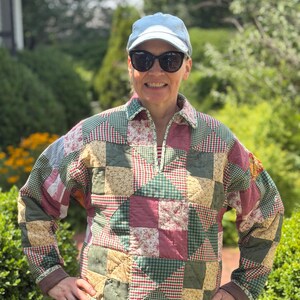 May include: A long-sleeve pullover jacket featuring a patchwork quilt design with various fabric squares in shades of green, red, and tan. The jacket has a collared neckline with a partial front zipper, long sleeves with solid brown cuffs, and a hem.