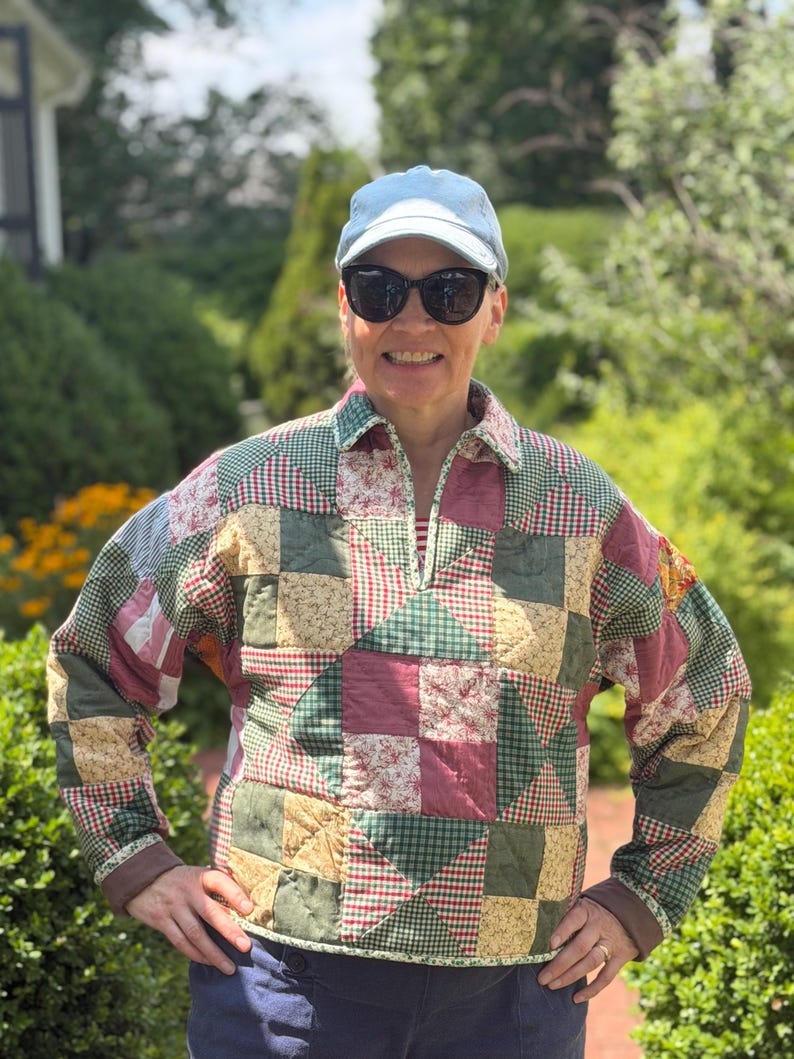 May include: A long-sleeve pullover jacket featuring a patchwork quilt design with various fabric squares in shades of green, red, and tan. The jacket has a collared neckline with a partial front zipper, long sleeves with solid brown cuffs, and a hem.