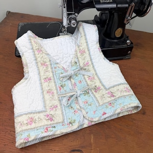 May include: A quilted vest with a floral pattern in pink, cream, and blue. The vest has a white background and is decorated with bows. A vintage black Singer sewing machine is in the background.