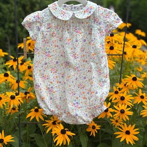 May include: A white short-sleeved romper featuring a delicate floral pattern in shades of purple, pink, and orange. The garment includes a rounded Peter Pan collar, vertical pleats across the chest, and elasticized leg openings for a gathered look.