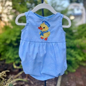 May include: A light blue baby romper with a small, repeating pattern. The romper features an embroidered yellow duck wearing a blue sailor hat and bow tie. The garment is sleeveless and hangs on a white hanger.