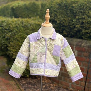 May include: A quilted jacket with a patchwork design in shades of lavender, green, and cream. The jacket features a button-up front and a collar. The fabric has floral and striped patterns.