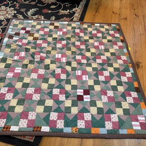 May include: A rectangular patchwork quilt featuring a repeating pattern of four-patch blocks and triangle squares. The fabric squares display various colors including dark green, maroon, cream, and tan, with small floral and checkered prints.