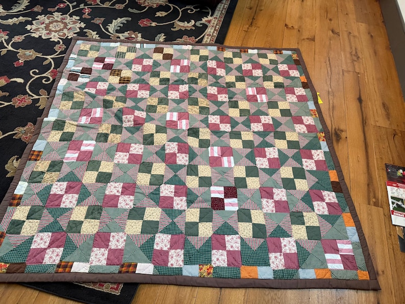 May include: A rectangular patchwork quilt featuring a repeating pattern of four-patch blocks and triangle squares. The fabric squares display various colors including dark green, maroon, cream, and tan, with small floral and checkered prints.