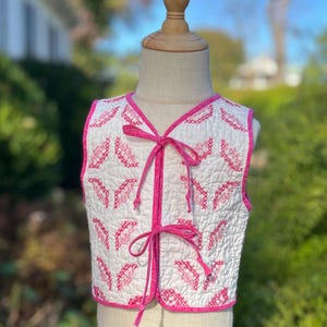 May include: White quilted vest with pink embroidered accents and trim. The vest features a V-neck and ties in the front with pink ribbons. The quilted pattern is a repeating design of pink embroidered shapes. The vest is displayed on a mannequin.