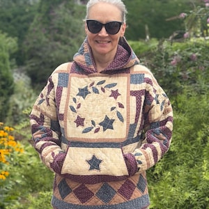 May include: A long-sleeve hooded pullover made from quilted fabric featuring a patchwork design in shades of navy blue, burgundy, and cream. The front displays a central star and leaf wreath motif, a large kangaroo pocket, and a single star applique.