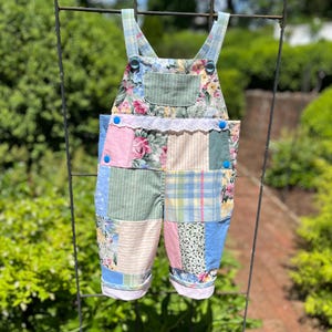 May include: A patchwork baby overall with floral, striped, and plaid patterns in pastel colors. The bib features a floral print and is trimmed with white lace. Blue buttons secure the straps and sides.