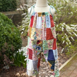 May include: A patchwork jumpsuit in a variety of colors and patterns, including floral, paisley, and solid fabrics. The jumpsuit has a halter-style top with light green ties and cropped pants. The garment is displayed on a mannequin.