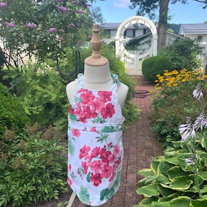 May include: A white apron with a vibrant floral pattern of pink and red flowers and green leaves. The apron is displayed on a wooden mannequin in a garden setting, showcasing its design and style. The apron has a pocket.