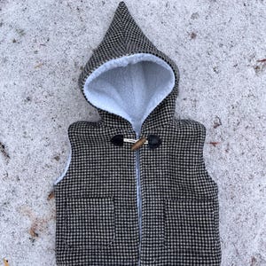 May include: A black and white checkered hooded vest with a toggle closure. The vest has a white, fluffy interior and two front pockets. The hood is pointed and lined with the same white material. The vest is designed for children.