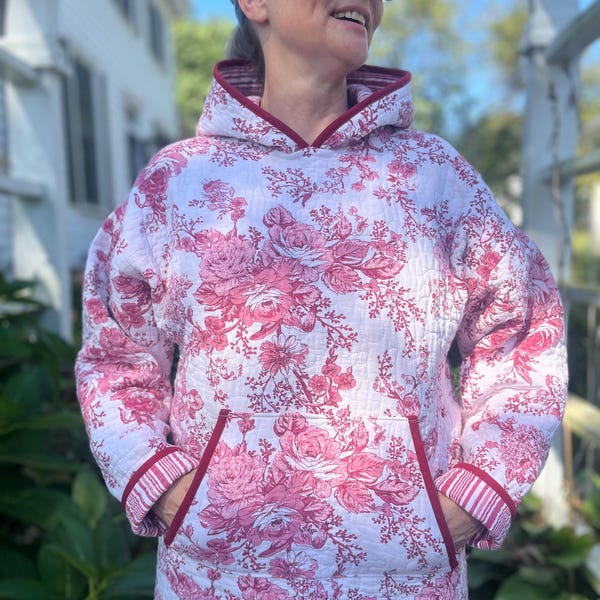 May include: A white and pink floral print hoodie with a kangaroo pocket and a hood. The hoodie has a red trim around the hood, pocket, and cuffs. The floral pattern features large roses and other flowers.