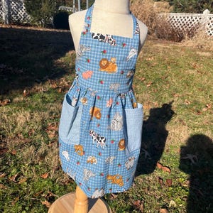 May include: A blue and white checkered apron with a variety of animal illustrations, including lions, cows, pigs, and elephants. The apron has two large pockets on the front.