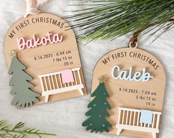 May include: Two arched wooden ornaments labelled My First Christmas. One features the name Dakota in pink with birth details 16.08.2025, 3.6 kg, 50.8 cm, a green tree, and a white crib with a pink blanket. The other says Caleb in blue with 19.09.2025, 2.6 kg, 48.3 cm, a green tree, and a white crib with a blue blanket.