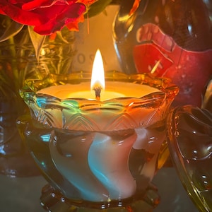 Könnte beinhalten: Ein gläserner Kerzenhalter mit einem wirbelnden, mehrfarbigen Design. Die Kerze ist angezündet und die Flamme wirft einen warmen Schein. Der Halter steht auf einem Tisch mit einer roten Rose und anderen Blumen im Hintergrund.