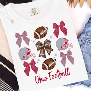May include: White t-shirt with a football theme. The shirt features illustrations of footballs, helmets, and bows in shades of brown, grey, and pink. The words "Ohio Football" are written in red script.