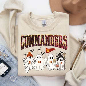 May include: A cream-colored sweatshirt with the word "COMMANDERS" in a glittery, maroon and gold font. Below the text are four cartoon ghosts with football jersey numbers and Halloween-themed accessories. A burgundy pennant with a white "W" is also present.