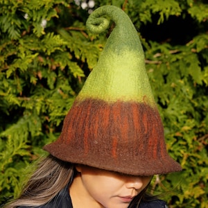 May include: A whimsical, handmade felt hat with a pointed, curled top. The hat transitions from a green tip to a light green midsection, and a brown base. The brim is wide, and the hat appears to be made of wool.
