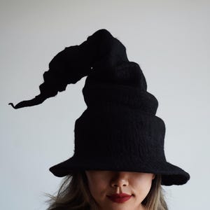 Op de afbeelding: Een zwarte heksenhoed met een brede rand en een hoge, spiraalvormige kroon. De hoed is gemaakt van een textuurrijk materiaal en staat tegen een effen witte achtergrond. De unieke vorm en donkere kleur van de hoed creëren een opvallend visueel effect.