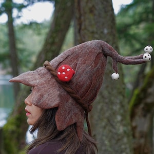 Puede incluir: Un sombrero de fieltro marrón caprichoso con un ala en forma de hoja y un motivo de seta. El sombrero presenta una seta roja con lunares blancos y un detalle de cuerda trenzada. Dos pequeños acentos blancos en forma de seta cuelgan de la punta del sombrero.