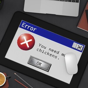 May include: A black rectangular mousepad with a computer error message that reads "You need more chickens." A white mouse, laptop, notebooks, pens, and a cup of coffee are also on the dark surface.