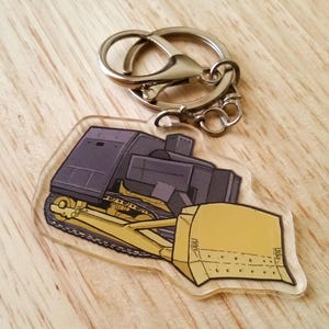 May include: A yellow and gray bulldozer keychain with a silver metal key ring. The bulldozer is facing left and has a large yellow blade.