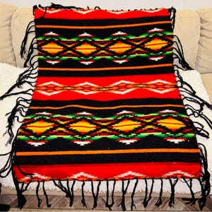 May include: A rectangular, woven blanket with a vibrant geometric pattern. The blanket features a red base with black stripes and diamond shapes in yellow, orange, and green. Black fringe trims the edges, adding texture and visual interest.