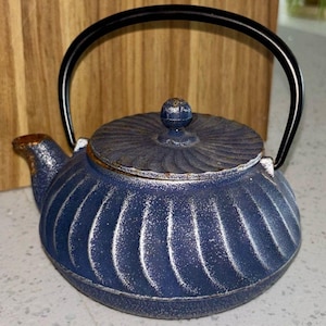 Puede incluir: Una tetera azul de hierro fundido con asa y tapa negras. La tetera tiene un diseño texturizado y acanalado con detalles blancos. La tapa está rematada con un pequeño pomo redondo. El pico tiene un acabado marrón. Un artículo de cocina decorativo.