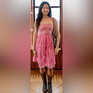 Puede incluir: Un vestido de verano rosa con un corpiño estampado y una falda fluida. El vestido se combina con botas de vaquero marrones. Se ve un bolso de hombro estampado. El fondo incluye una ventana y molduras de madera.