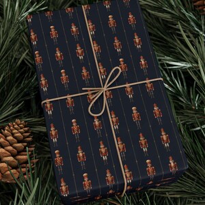 May include: A wrapped gift with a navy blue background and a repeating pattern of toy soldiers in red, gold, and brown. The gift is tied with twine and rests on a bed of pine needles and a pine cone.