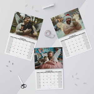 May include: A set of three 2024 calendars featuring a man with angel wings and a cat. The calendars are open to the months of October, November, and December. The man is wearing a tutu in each image.