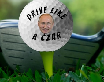 Może przedstawiać: Piłka golfowa na zielonym tee z napisem "DRIVE LIKE A CZAR" i twarzą nałożoną na piłkę. Kij golfowy jest w tle, z zieloną trawą.
