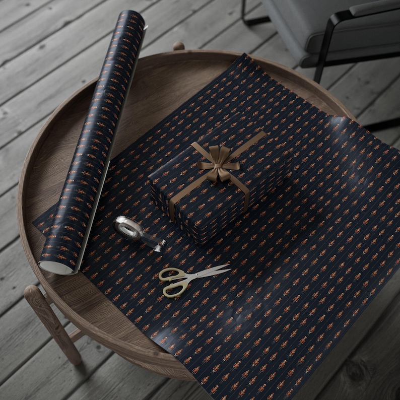 Puede incluir: Papel de regalo azul marino con un patr&oacute;n naranja repetido, un regalo envuelto, tijeras y un dispensador de cinta adhesiva sobre una mesa de madera. El regalo est&aacute; atado con una cinta marr&oacute;n.