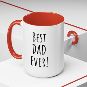 Op de afbeelding: Witte keramische mok met een rode rand en handvat. De mok heeft de tekst "Best Dad Ever!" in zwart gedrukt.