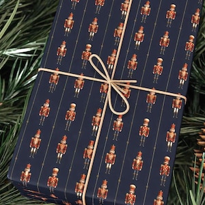 May include: A wrapped gift with navy blue wrapping paper featuring a pattern of toy soldiers in red and gold. The gift is tied with a gold ribbon and bow, and is surrounded by pine needles.