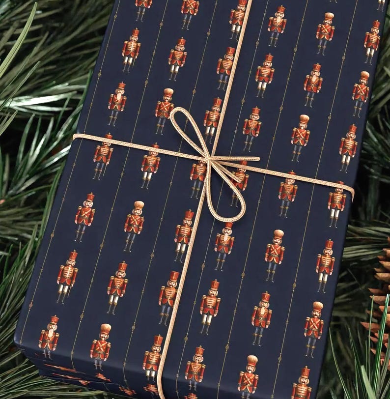 May include: A wrapped gift with navy blue wrapping paper featuring a pattern of toy soldiers in red and gold. The gift is tied with a gold ribbon and bow, and is surrounded by pine needles.
