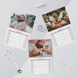 May include: Three spiral-bound calendars featuring whimsical photos of a man with a beard and angel wings, posing with cats. The calendars are open to January, February, and March 2024, with a white background.