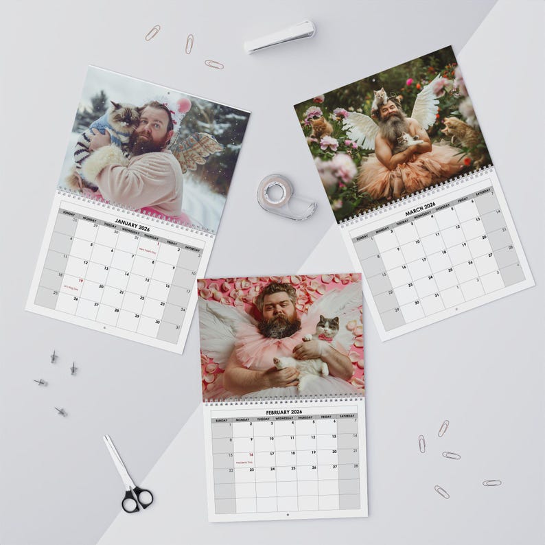 May include: Three spiral-bound calendars featuring whimsical photos of a man with a beard and angel wings, posing with cats. The calendars are open to January, February, and March 2024, with a white background.