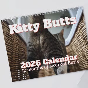 Può includere: Un calendario a spirale intitolato "Kitty Butts 2026 Calendar". Il calendario presenta il posteriore di un gatto tigrato in un cesto di vimini. È visibile anche il testo "12 months of sexy cat Butts".